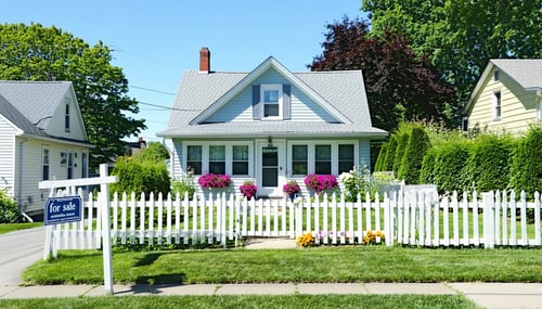 A cozy suburban home stands proudly against a backdrop of lush greenery framed by a white picket fence The house features a charming faade with light blue siding and crisp white trim adorned with blooming flower boxes under the windows A wellmanicure