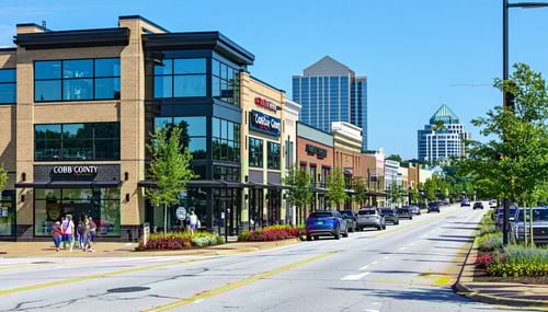 The image depicts a bustling commercial district in Cobb County showcasing a variety of commercial properties ranging from modern office buildings to retail storefronts The buildings are characterized by large glass windows reflecting the blue sky vi-3