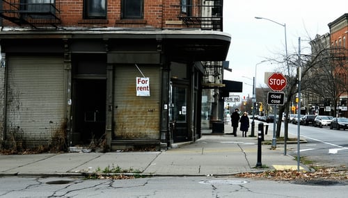 The image depicts a oncebustling street now lined with shuttered storefronts their windows dark and covered in dust A For Rent sign hangs in the window of a former caf while a nearby boutique displays a faded Closed sign The pavement is cracked and w