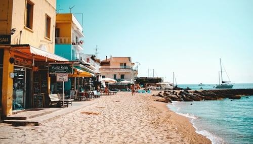 The image depicts a picturesque coastal town once bustling with tourists now showing signs of decline The vibrant beachfront normally filled with sunbathers and families is nearly empty with only a few scattered beach towels and abandoned umbrellas T