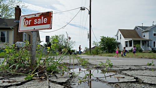 The image depicts a rundown suburban neighborhood with cracked pavement and overgrown weeds sprouting through the gaps In the foreground a faded For Sale sign leans at an angle in a yard with unkempt grass An old rusted power line hangs low and a nea