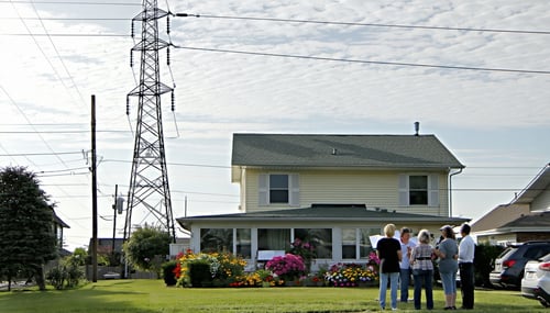 The image depicts a suburban landscape where a large metallic telecommunication tower looms prominently in the background casting a long shadow over a wellmaintained residential property The house a modest twostory structure with a neatly trimmed law